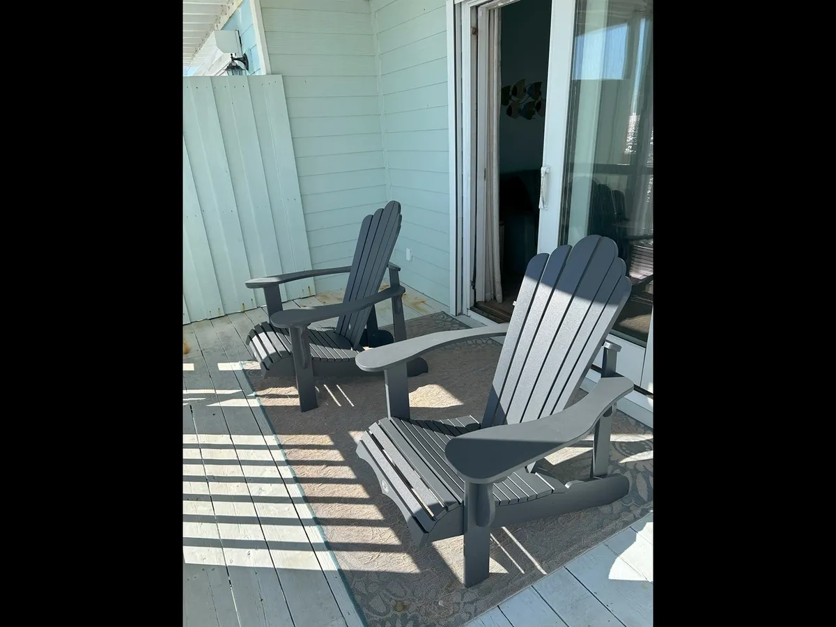 An inviting exterior shot featuring two Adirondack chairs placed on an outdoor rug by a sliding glass door, creating a cozy space for relaxing and soaking in the sea air. The arrangement invites guests to share stories, enjoy the view, or simply unwind together.