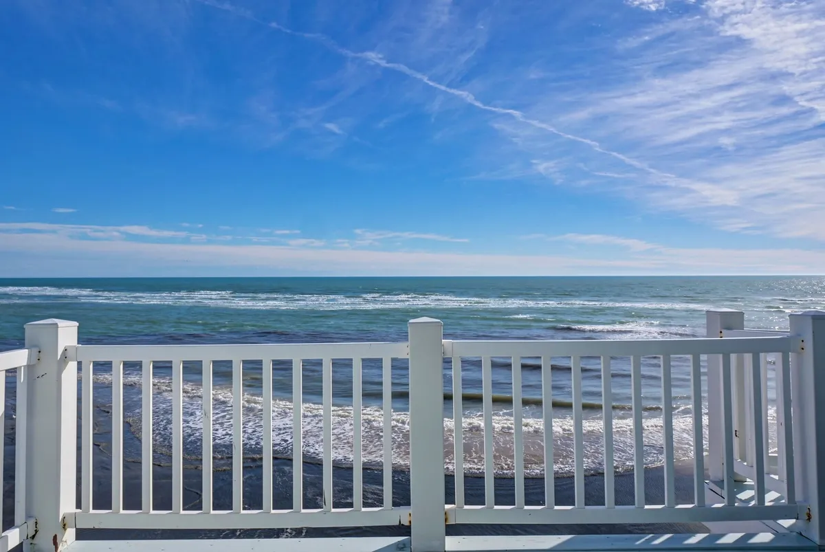 A tranquil view of the ocean framed by a simple wooden fence, with waves rolling gently onto the shore. The sky glows softly, evoking the peaceful atmosphere of a sunset or early morning, ideal for unwinding or taking a reflective walk along the beach.