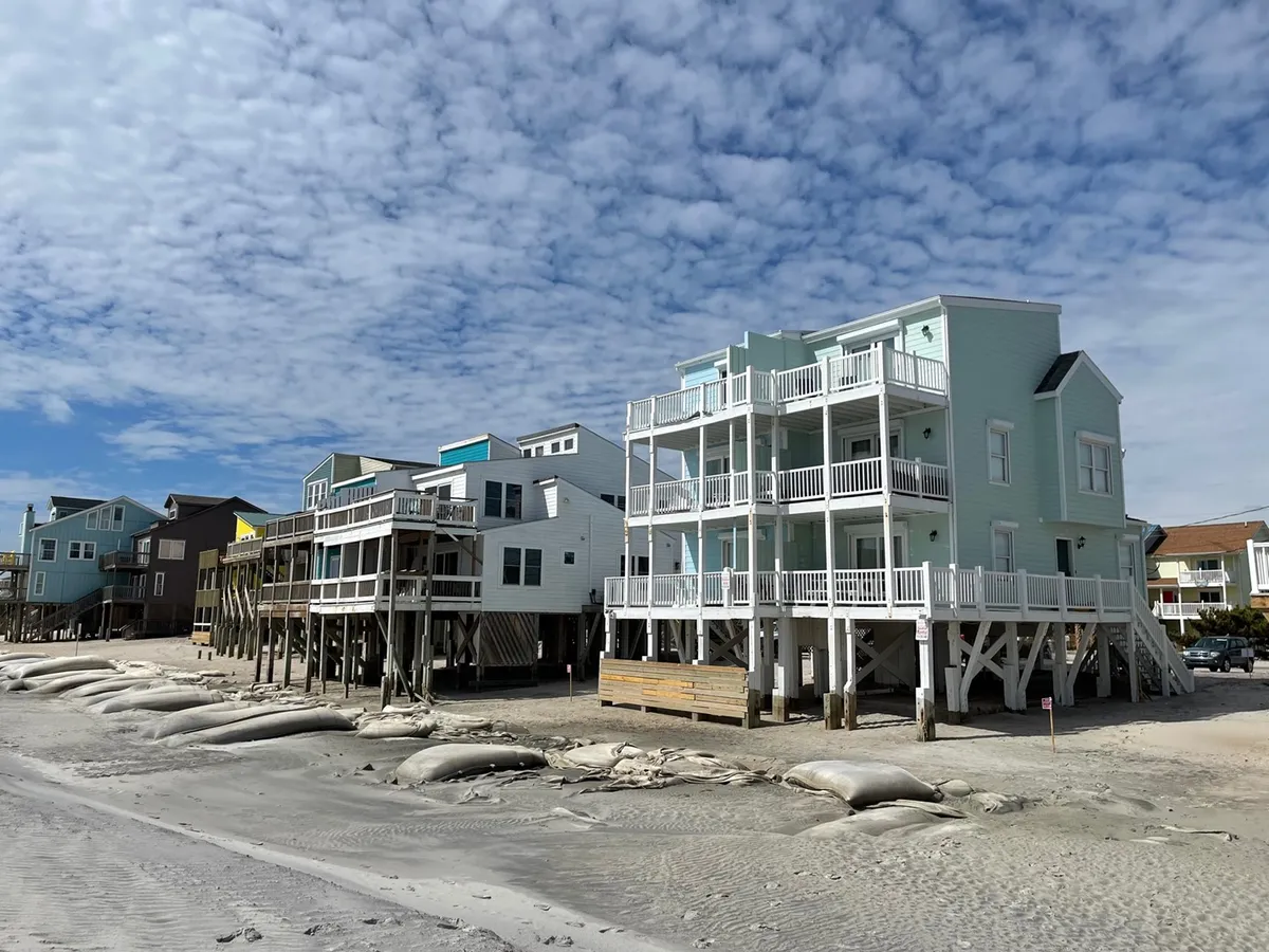 A row of colorful beach houses lines the sandy shore, with sandbags protecting the coastline and an expansive sky dotted with gentle clouds overhead. People can be seen walking along the beach and enjoying the view from their balconies, giving the scene a cheerful, active vibe.