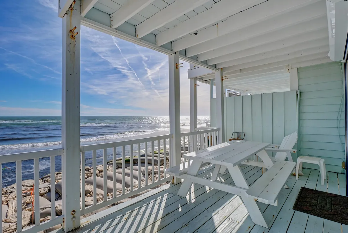 A serene deck area with a wooden picnic table set against a backdrop of sweeping ocean views. The deck is partially covered with wooden beams, creating a welcoming spot to relax, dine, or watch the waves. The tranquil setting suggests private outdoor enjoyment directly facing the sea.