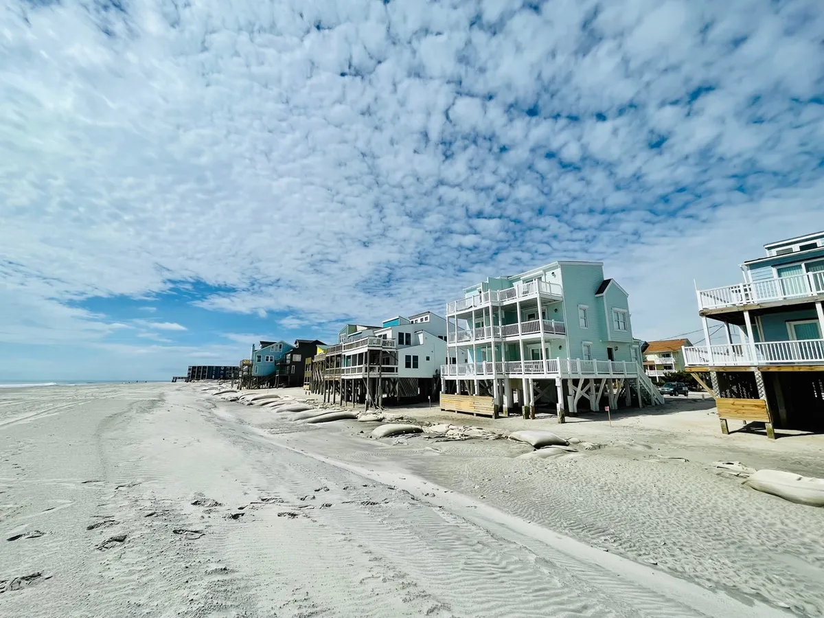A serene beachfront scene showcasing sand dunes in the foreground, rows of beachfront houses, and a soft, cloudy sky. The inviting stretch of sand suggests opportunities for peaceful walks and quiet moments together.
