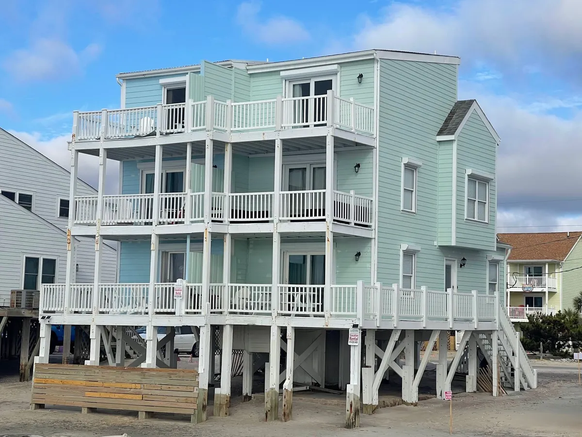 A multi-level light blue beach house sits elevated on a stilted foundation, with generous balconies at each level. The decks are set up for outdoor dining and relaxation, facing directly toward the ocean, making it an inviting spot to enjoy coastal breezes with a pet.