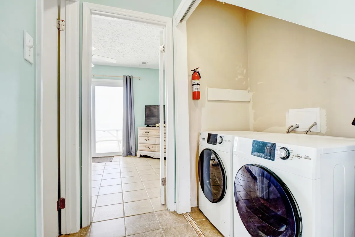 A bright, practical laundry area featuring a washer and dryer tucked neatly into a corner, with a doorway leading to another part of the home. The space feels fresh and welcoming, indicating convenience and ease for guests managing sandy towels and swimsuits after a day at the beach.