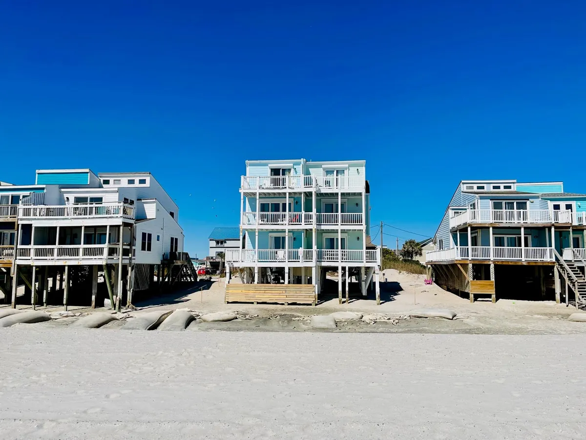 This bright exterior captures three beach houses, a sandy area perfect for games or sunbathing, and a clear blue sky. The lively, open space is well-suited for group activities like volleyball, frisbee, or water sports, which are mentioned as group-friendly activities.