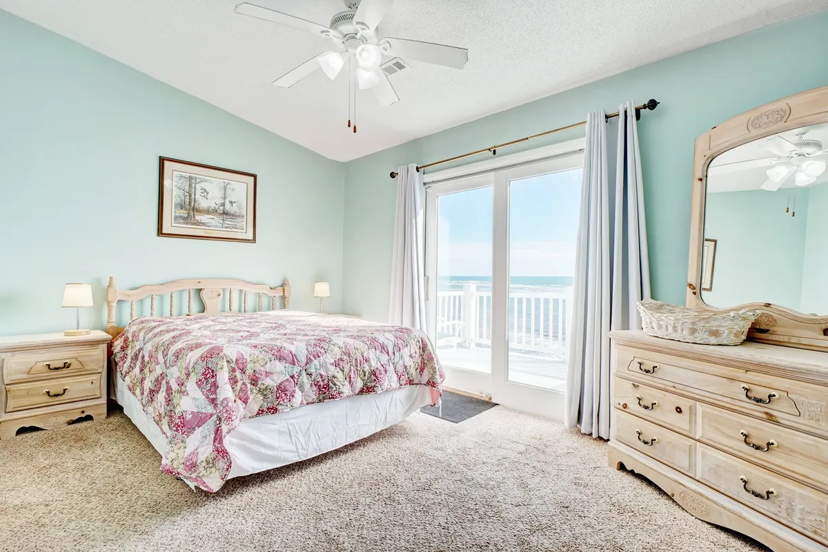 A serene bedroom featuring a bed dressed in a floral quilt, a large sliding door opening to reveal stunning ocean views, and a dresser with a mirror. The room is bright and airy, emphasizing tranquility and a direct connection to the outdoors, perfect for unwinding and taking in the coastal scenery.