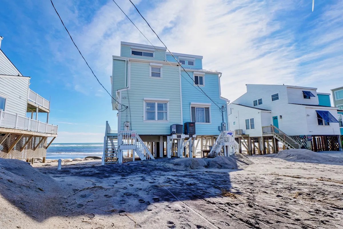 A welcoming exterior view of a beach house with a spacious sand area in front, surrounded by neighboring homes and with the ocean visible beyond. The deck offers ample space for outdoor relaxation, morning coffee, or family meals with a scenic ocean backdrop.