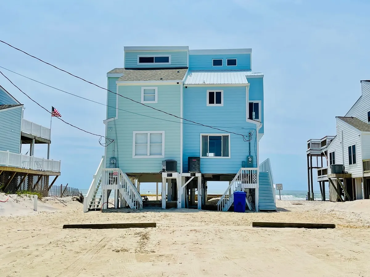 An inviting exterior view of the beach house with stairs leading up to the main entrance and the sandy beach area surrounding it. The house features decks overlooking the ocean, capturing the connection between indoor living and immediate beach access.