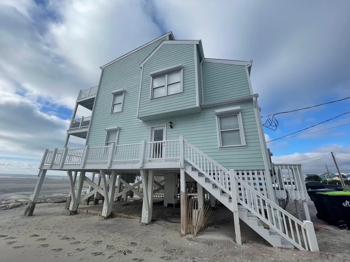 This exterior image shows a beach house structure with stairs leading up to a welcoming porch, set against a backdrop of the surrounding beach area. The inviting deck area, elevated entry, and expansive space suggest a perfect spot for group gatherings and enjoying the ocean breeze.