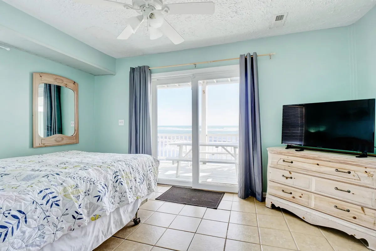 A master suite with a bed adorned in floral bedding, a large window offering unobstructed ocean views, a television set atop a dresser, and a mirror on the wall. The ambiance is tranquil and luxurious, with natural light pouring in to highlight panoramic Atlantic vistas.