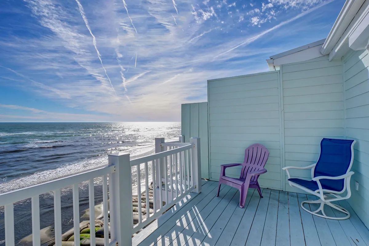 A serene view from the oceanfront deck with deck chairs inviting you to relax while gazing at expansive blue skies and the shimmering ocean. The scene captures the seamless transition from indoor social spaces to outdoor relaxation, echoing the ‘heart of the home’ flowing directly onto the deck.