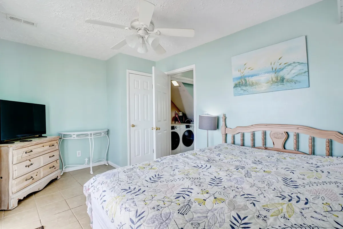 A peaceful bedroom featuring a bed with floral bedding, a TV, a dresser, and tasteful artwork on the wall. Sunlight streams softly into the room, highlighting the inviting atmosphere ideal for relaxation after a day at the beach.